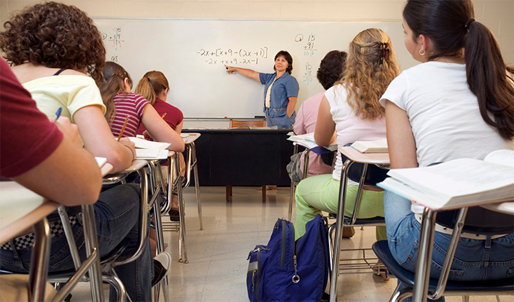 Photo of teenage students in a classroom looking at a whiteboard with a teacher pointing to a math equation.