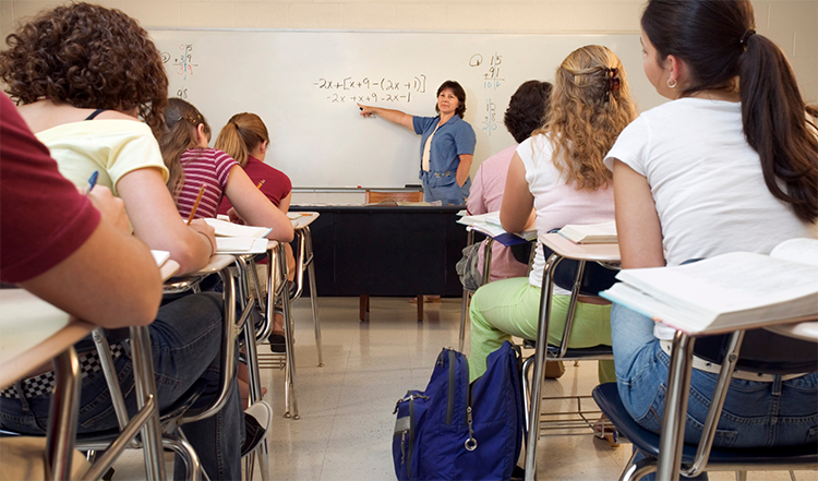 Photo of teenage students in a classroom looking at a whiteboard with a teacher pointing to a math equation. 